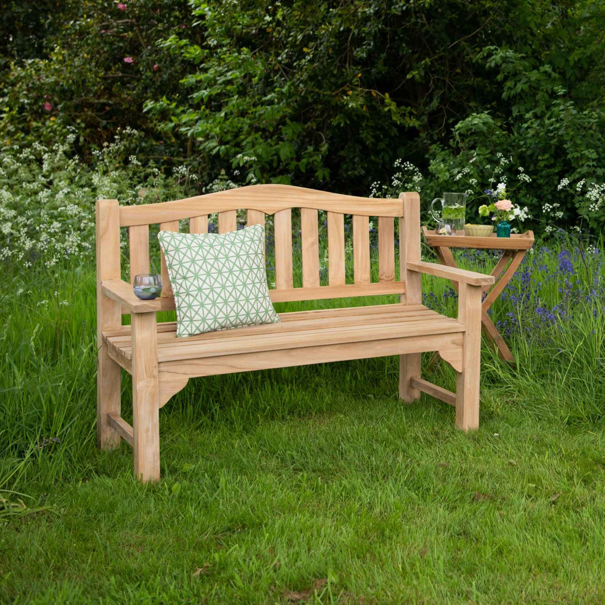 Teak bench with curved slatted backrest and slatted seat, with green patterned cushion, next to folding teak side table with flowers and jug, in outdoor setting