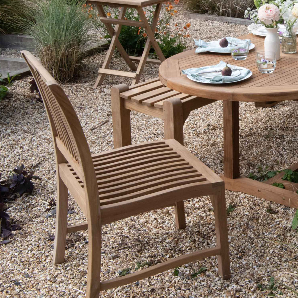 
                  
                    Teak dining chair and teak bench, next to  oval teak dining table with place settings and flowers, on gravel patio with folding teak table in background. 
                  
                
