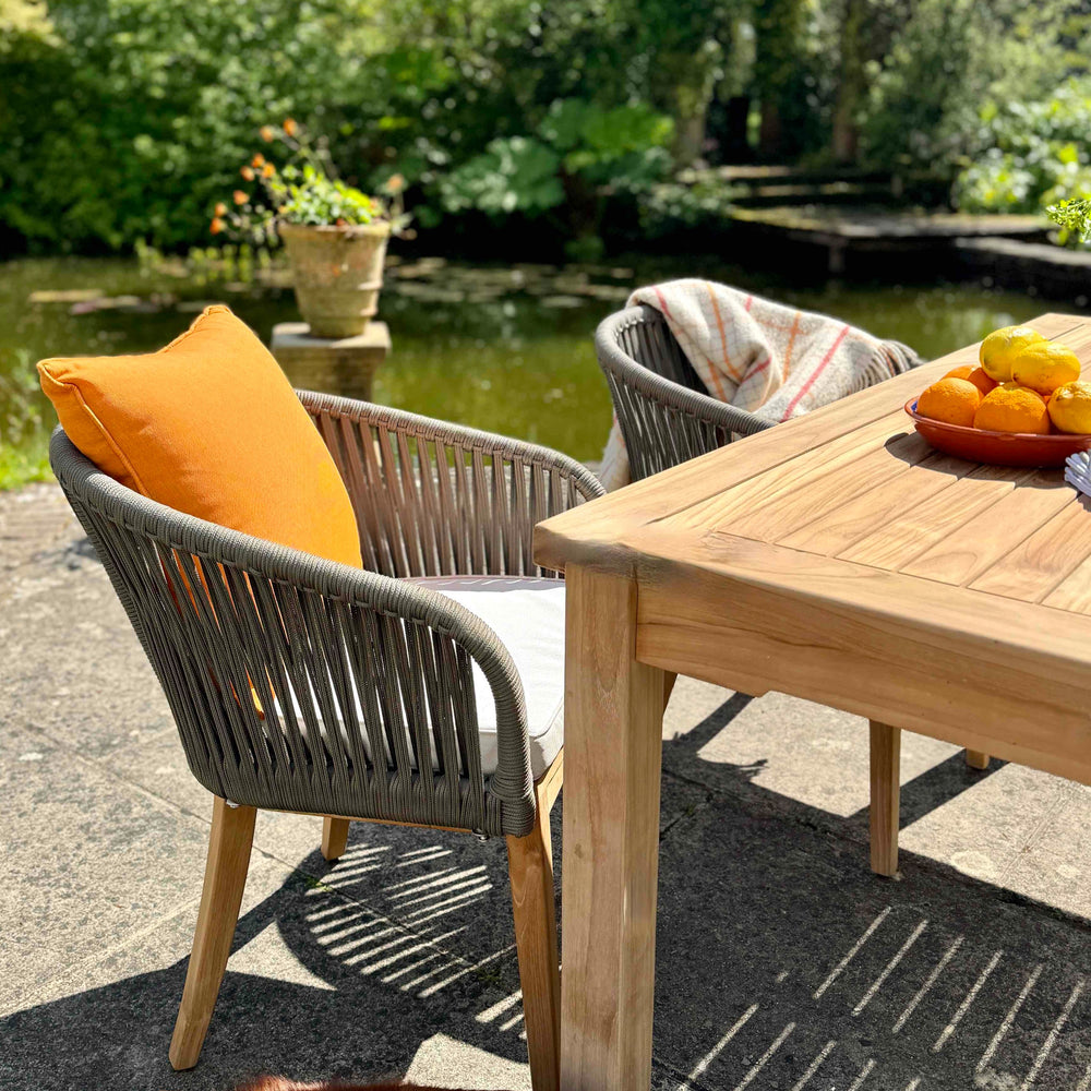 
                  
                    Two teak and rope outdoor dining chairs with orange scatter cushion and cream seat pad, next to teak dining table, on sunny patio with lake and trees in background
                  
                