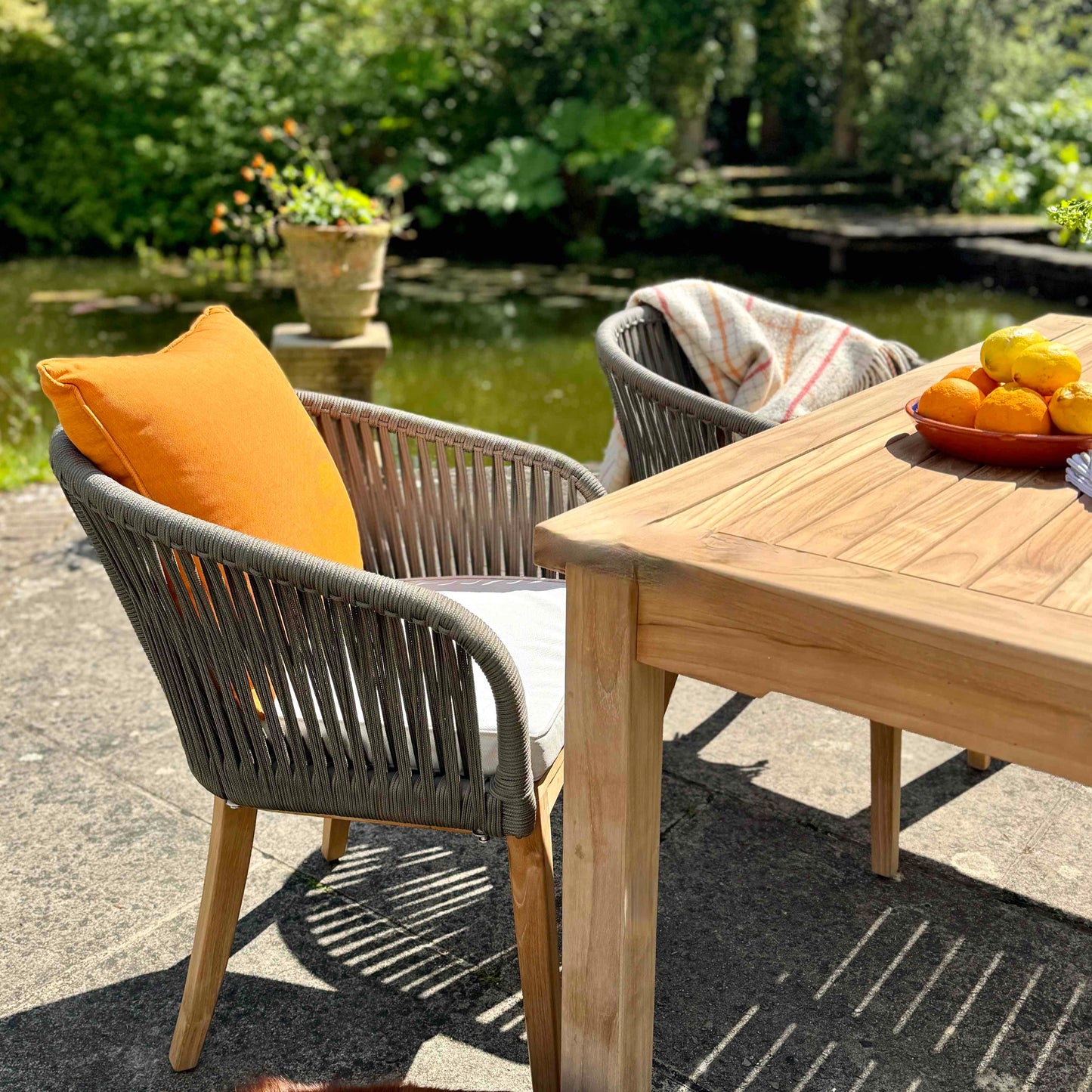 Two teak and rope outdoor dining chairs with orange scatter cushion and cream seat pad, next to teak dining table, on sunny patio with lake and trees in background