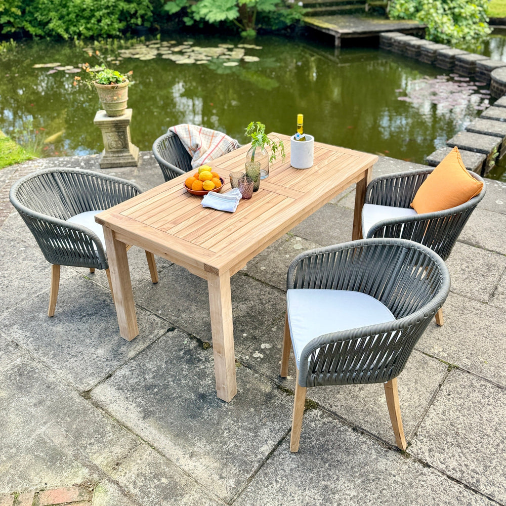 
                  
                    Teak rectangular dining table, with dish of oranges and plant, with four teak and rope dining chairs around, with matching cushions. A pond is in the background.
                  
                