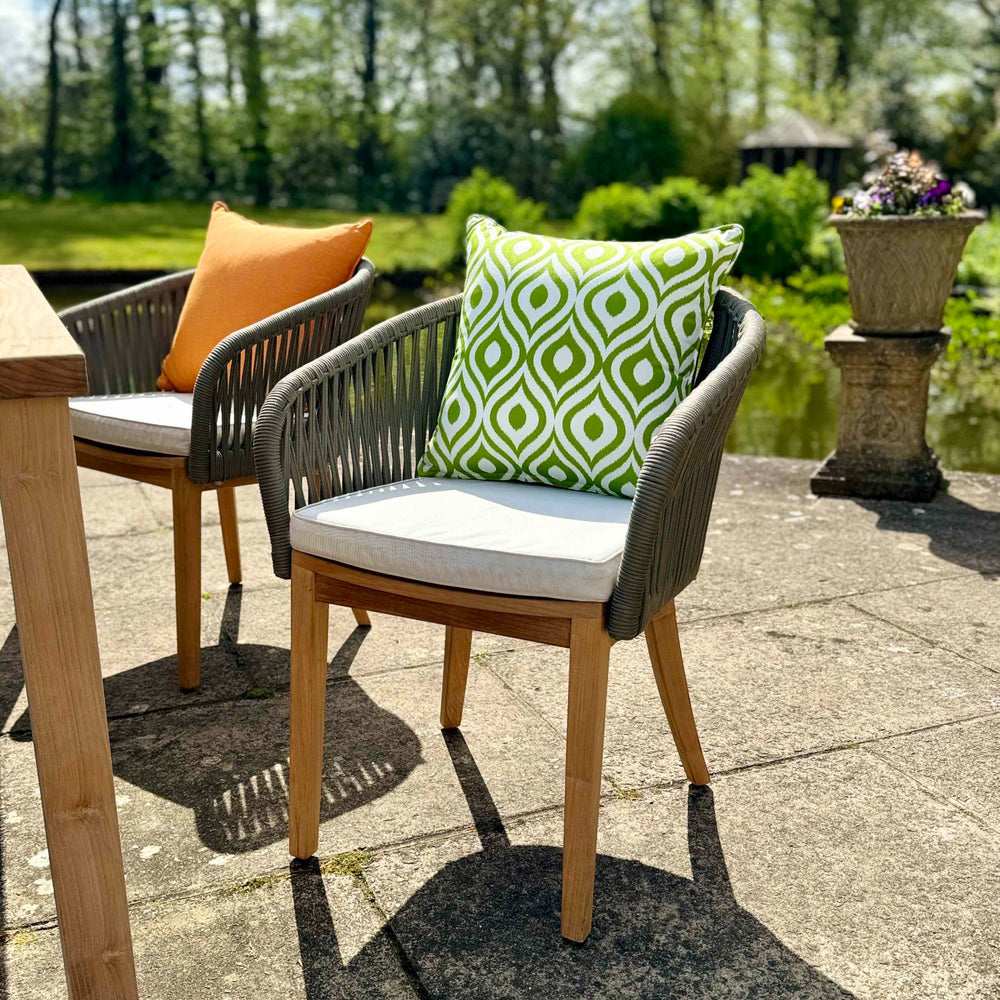 
                  
                    two teak outdoor dining chairs with rope backrests and armrests, with cream coloured seat cushions and green patterned and orange scatter cushions, on stone patio 
                  
                