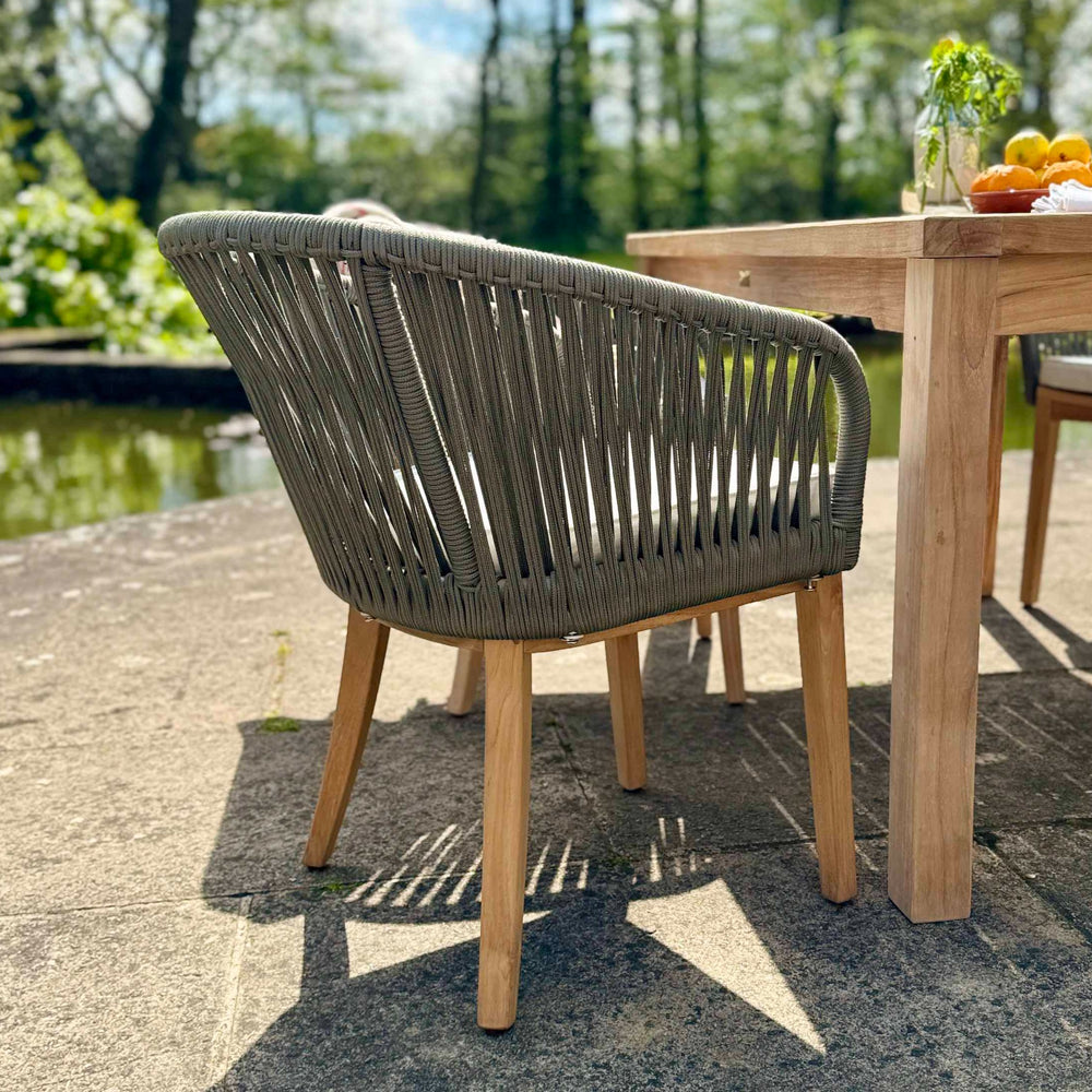 
                  
                    Back view of teak and rope outdoor dining chair with corner of teak dining table on which is bowl of fruit and vase of greenery. The chair is on stone patio with a pond behind. 
                  
                