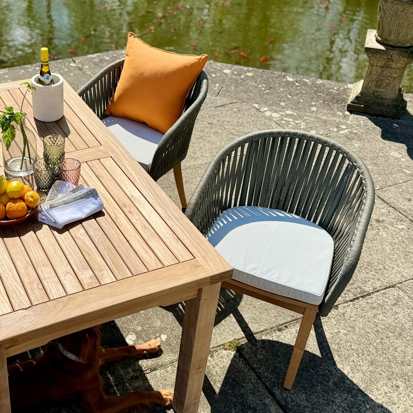 View from above of two teak and rope chairs with cream seat pads, on stone patio. There is a teak dining table with wine cooler and glasses. Behind is a pond and stone pedestal