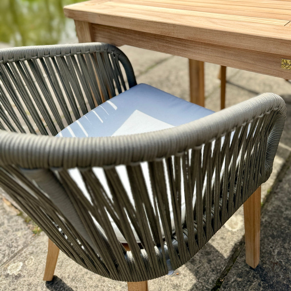 
                  
                    Close up of single teak and rope outdoor dining chair with cream cushion, pushed up against teak rectangular dining table. 
                  
                