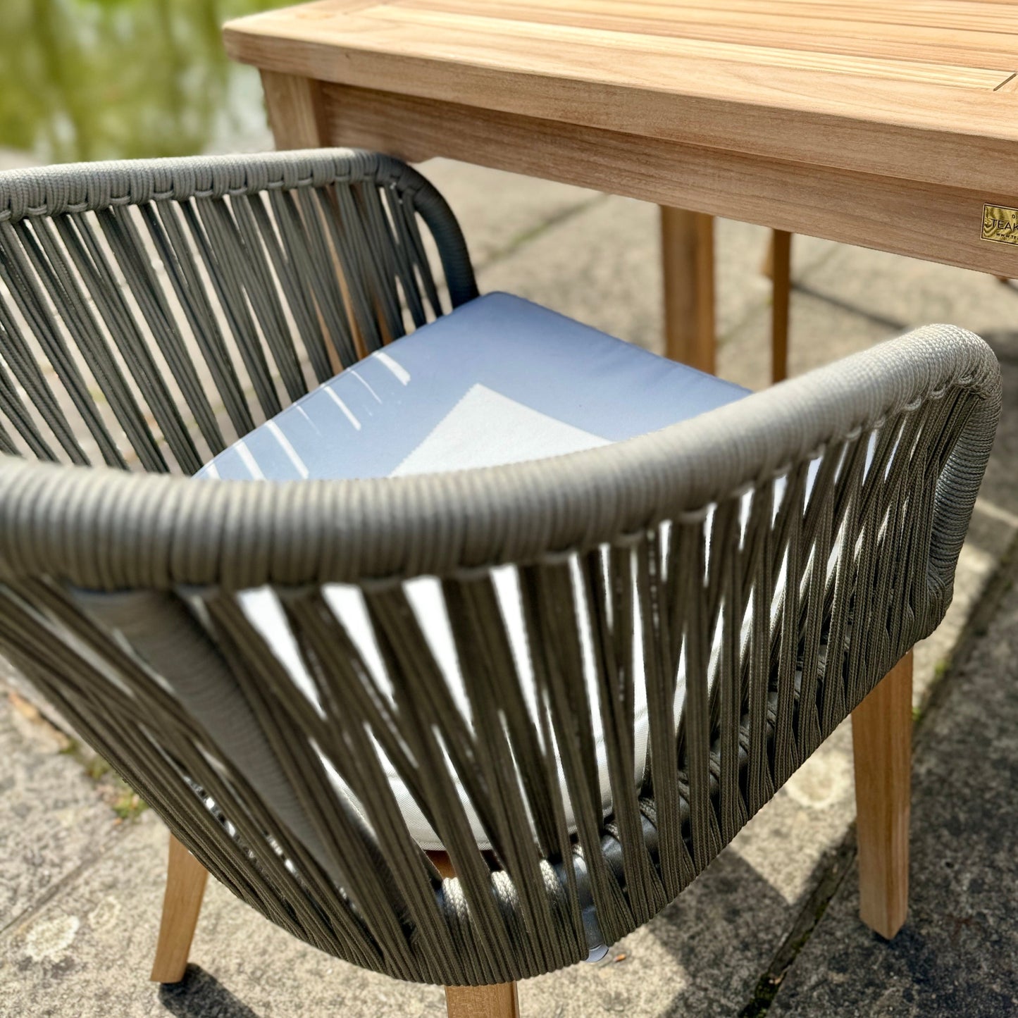 Close up of single teak and rope outdoor dining chair with cream cushion, pushed up against teak rectangular dining table. 