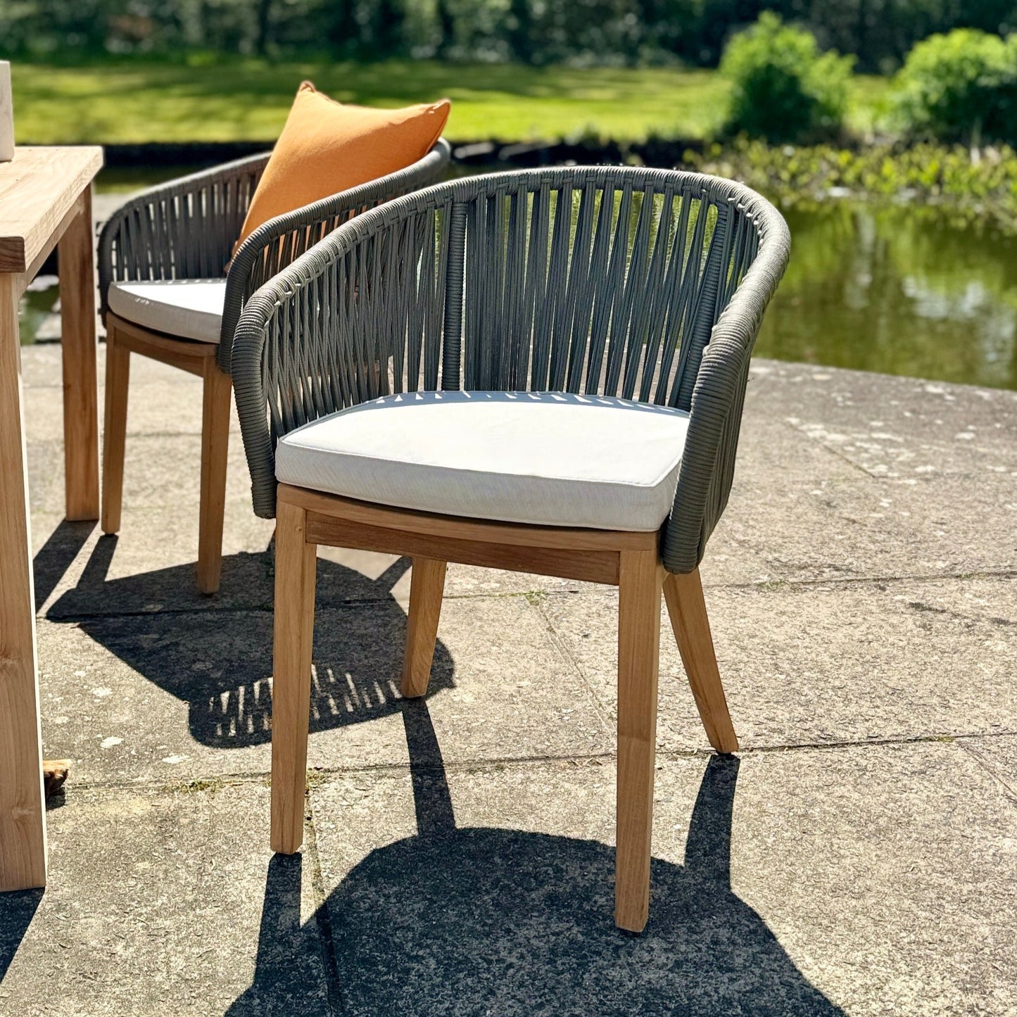 Two teak and rope chairs with cream seat pads, on stone patio, one with orange cushion. There is a teak dining table next to the chairs and a pond behind.  
