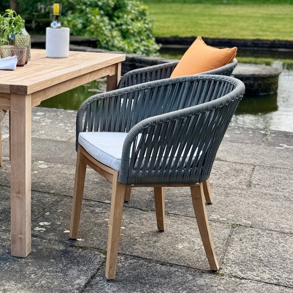 
                  
                    A teak and rope chair with solid teak base and legs, and backrest wrapped in grey rope weave and a pale grey seat pad, placed outdoors next to a teak dining table.
                  
                