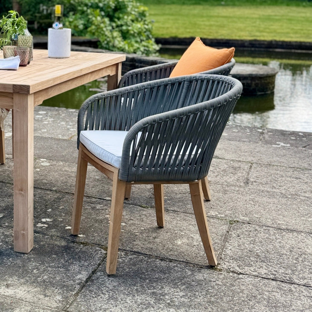
                  
                    End of teak rectangular dining table with teak chairs, with rope backrests and blue cushions, on stone patio with a pond in background
                  
                