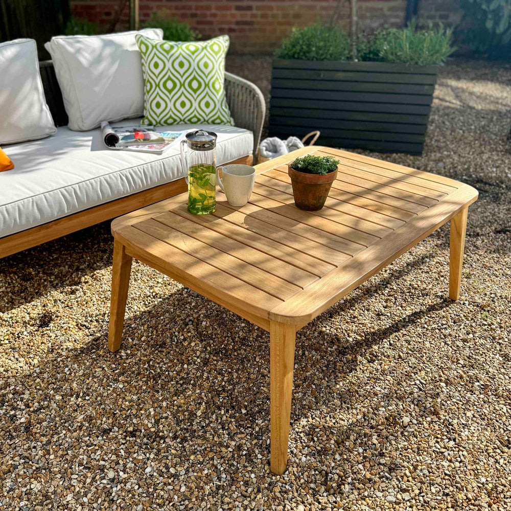 
                  
                    Teak outdoor sofa with white cushions on gravel with teak coffee table in front. Planters behind. 
                  
                