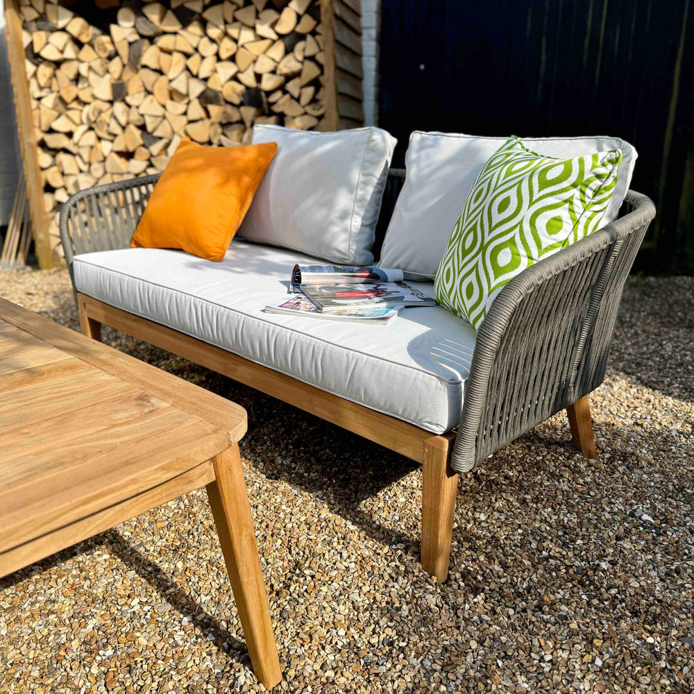 
                  
                    Two seater outdoor sofa made of teak base and rope sides and backrest with white seat and back cushions. In front is teak table.Logs piled in background. 
                  
                