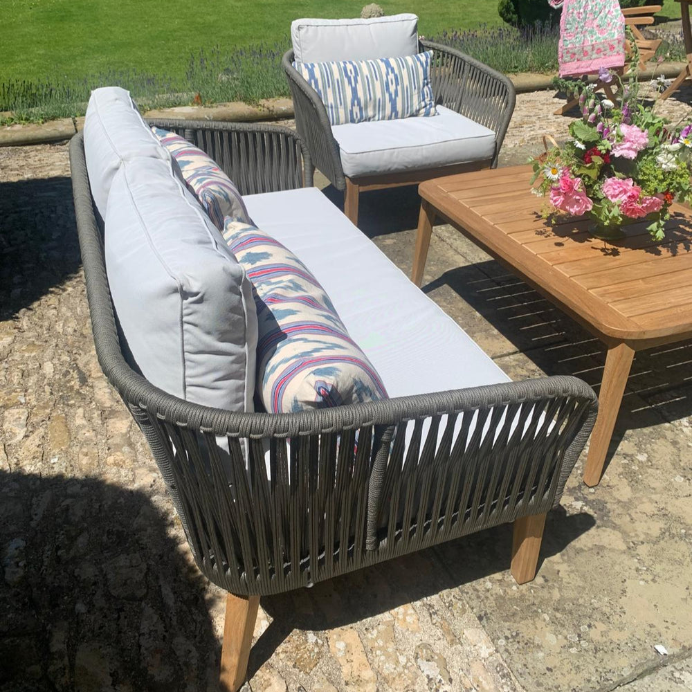 
                  
                    Outdoor sofa and chair set with teak legs and rope backrest and armmrests,with white and colourful cushions. In front is a teak table, in a garden setting.
                  
                