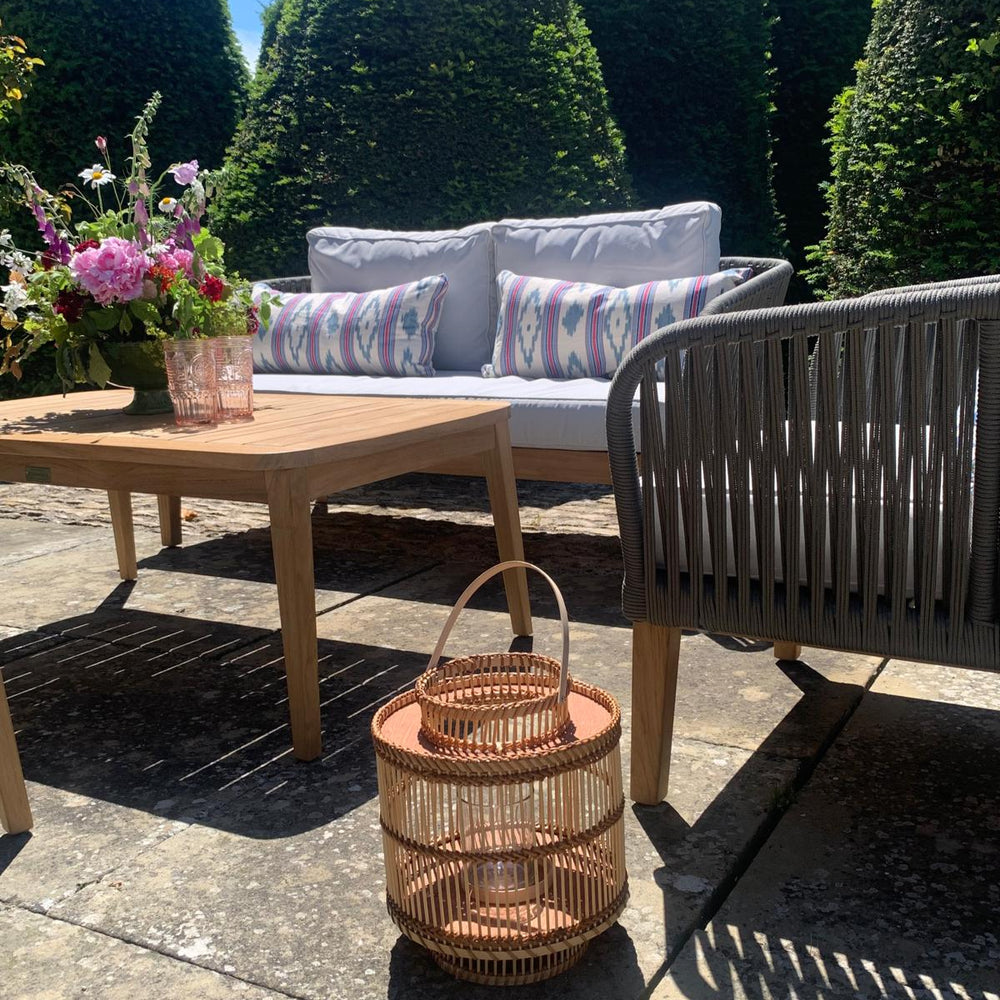 
                  
                    Outdoor sofa and chair set with teak legs and rope backrest and armmrests,with white and colourful cushions. In front is a teak table, and wooden lantern, in a garden setting.
                  
                