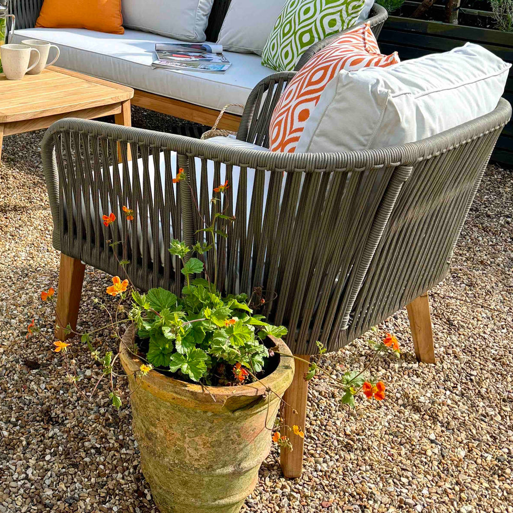 
                  
                    Teak outdoor dining chair with rope backrest and arms, with patterned scatter cushion and cream coloured back and seat pads, sitting next to teak and rope outdoor sofa and teak coffee table, on gravel patio.
                  
                