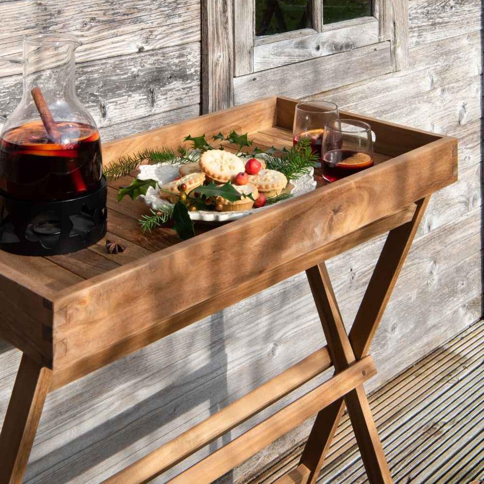 A butler's tray made of teak wood on a stand, displayed with drinks and food on top, placed on a wooden deck outdoors.