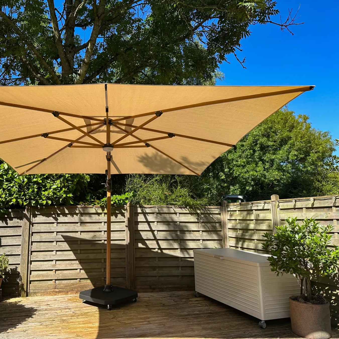 Beige patio umbrella on a wooden deck with greenery and a wooden fence in the background.