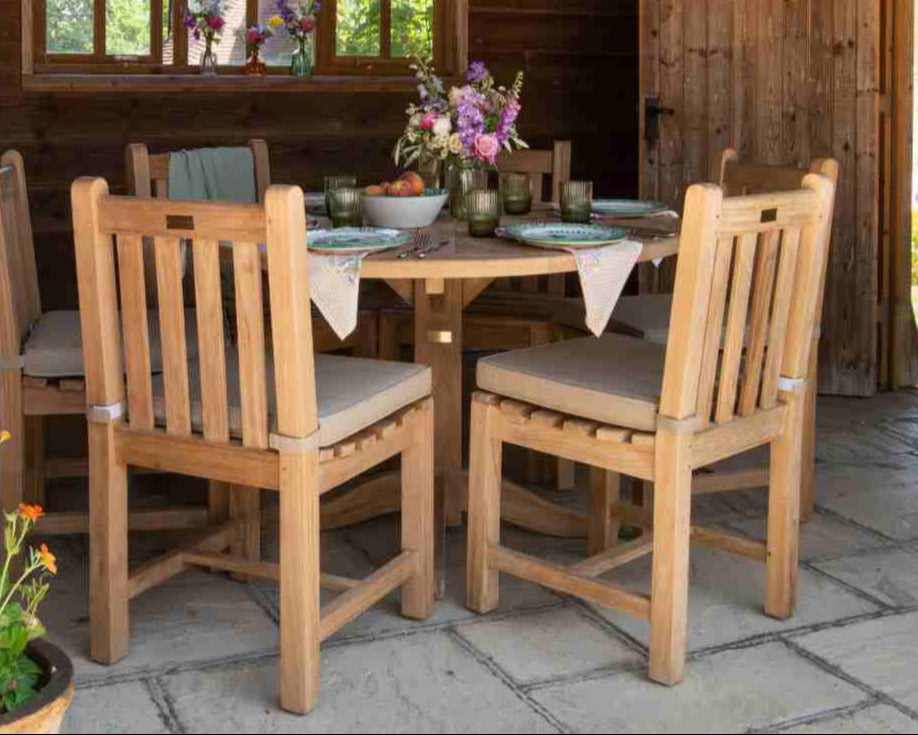 
                  
                    Teak dining table with teak chairs and a floral arrangement in an outdoor setting.
                  
                