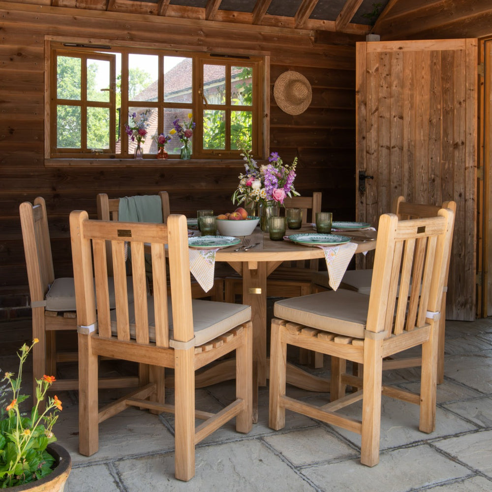 Round teak dining table set with flowers and place settings, with teak dining chairs with oatmeal coloured cushions, on stone patio with wooden wall and windows behind. 
