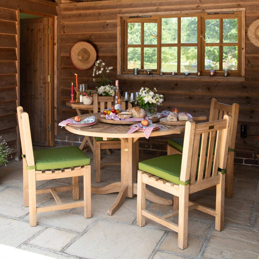 
                  
                    Round teak dining table, set with place settings, flowers, bread board and wine, with teak, green-cushioned chairs around and wooden wall and windows in the background. 
                  
                
