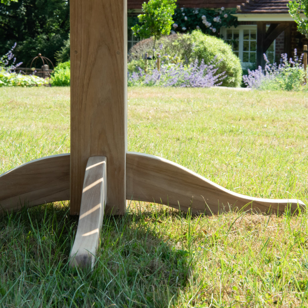 Close up of pedestal leg of round teak table, sitting on lawn with flowerbeds and building in background. 