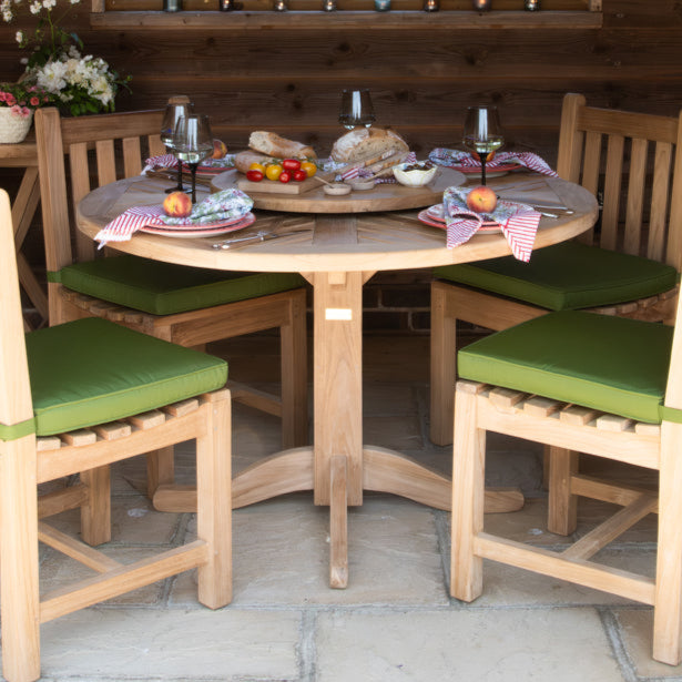 A teak round dining table set with green cushioned chairs and a circular dining setting in the background, placed on a patio.