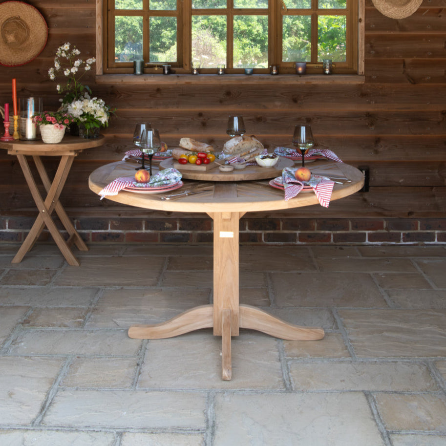 Round teak dining table set with glasses and place settings, on paved patio with wooden wall and window in background