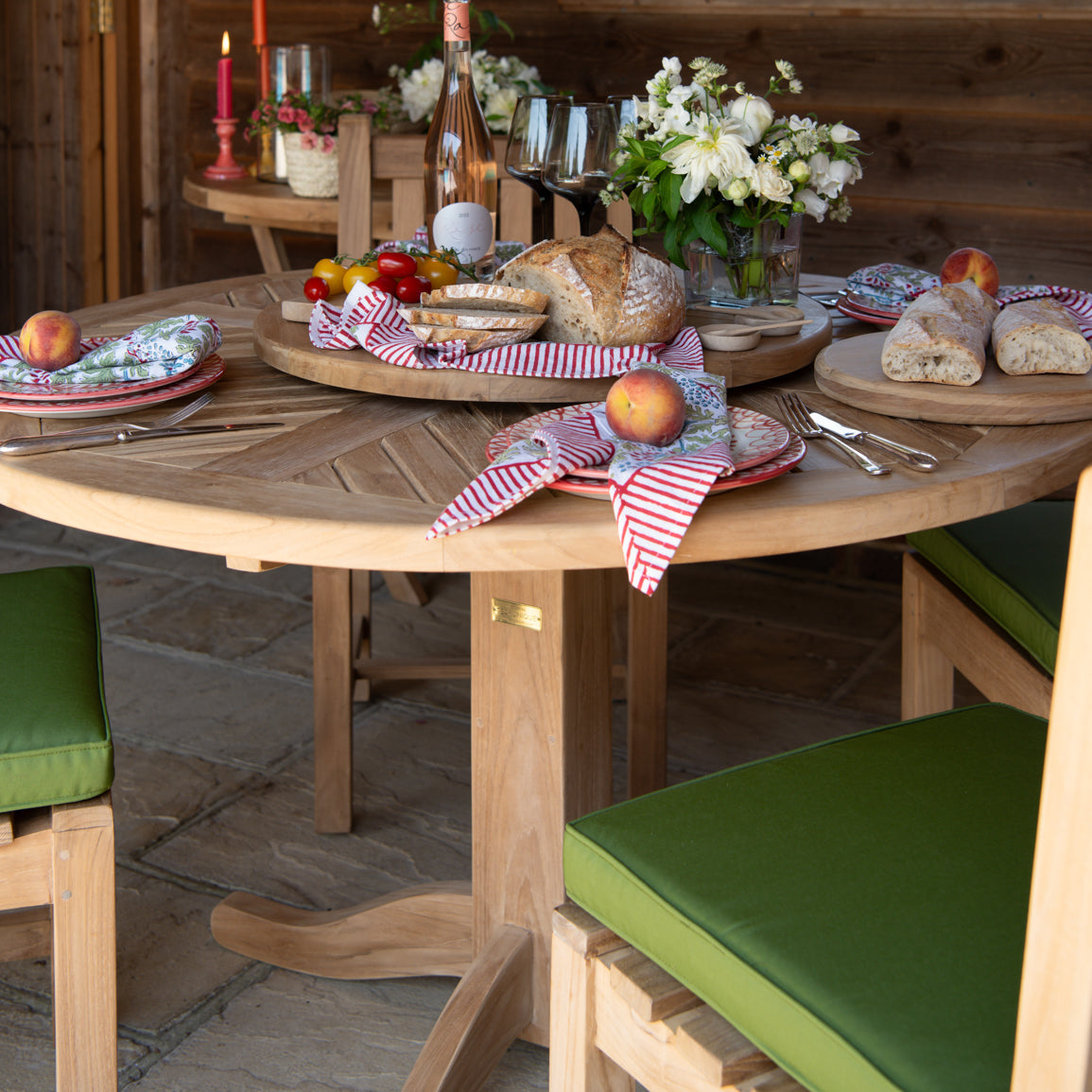Round teak dining table set with place settings, wine, bread board, flowers and glasses, with green cushioned chairs set around.  