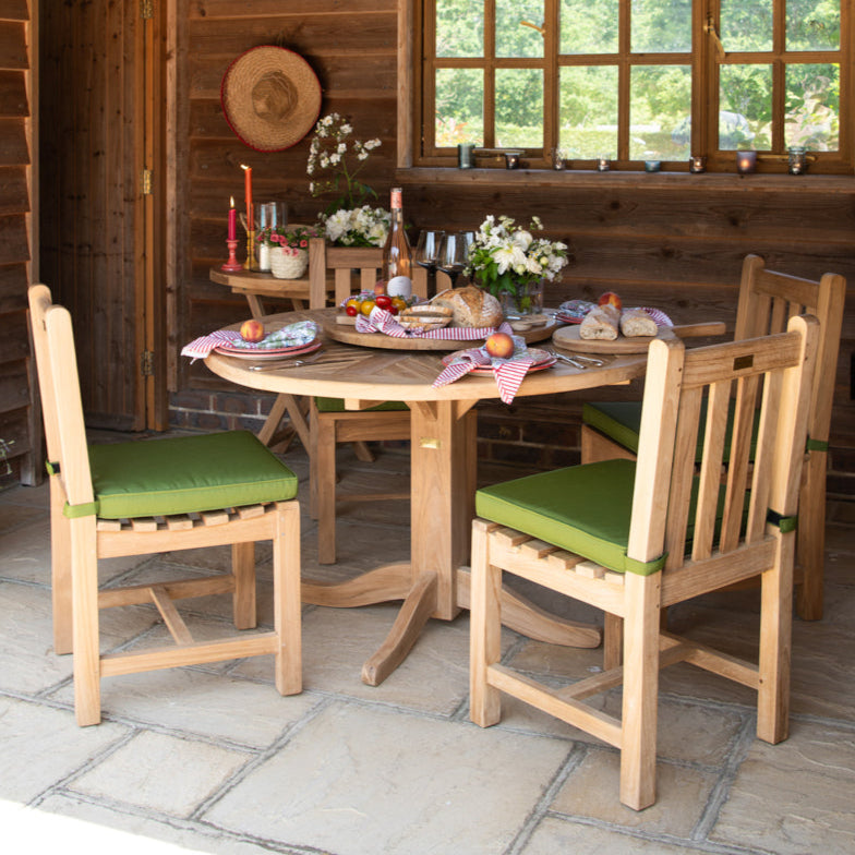 Round teak dining table, set with bread board, wine and place settings, with wooden wall and window in background, and green cushioned teak chairs around.  
