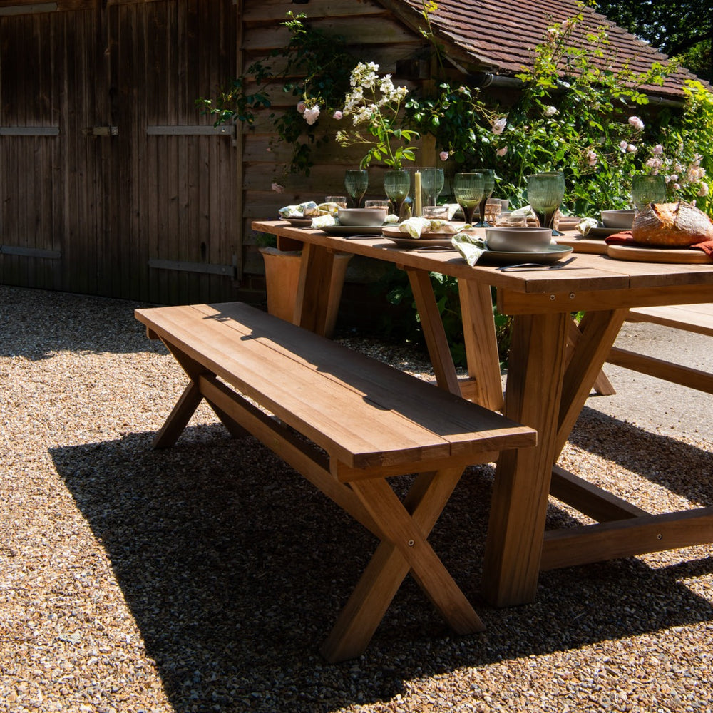 
                  
                    Teak rectangular outdoor dining table, set with plates, flowers and glasses with teak bench, on gravel patio
                  
                