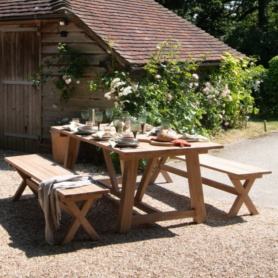 Teak rectangular outdoor dining table and two teak benches, set with place settings and glasses, with roses and wooden building in background