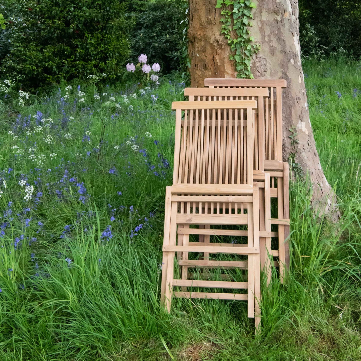 three teak folding dining chairs, in folded position, stacked against tree, in garden setting with wild floweres.