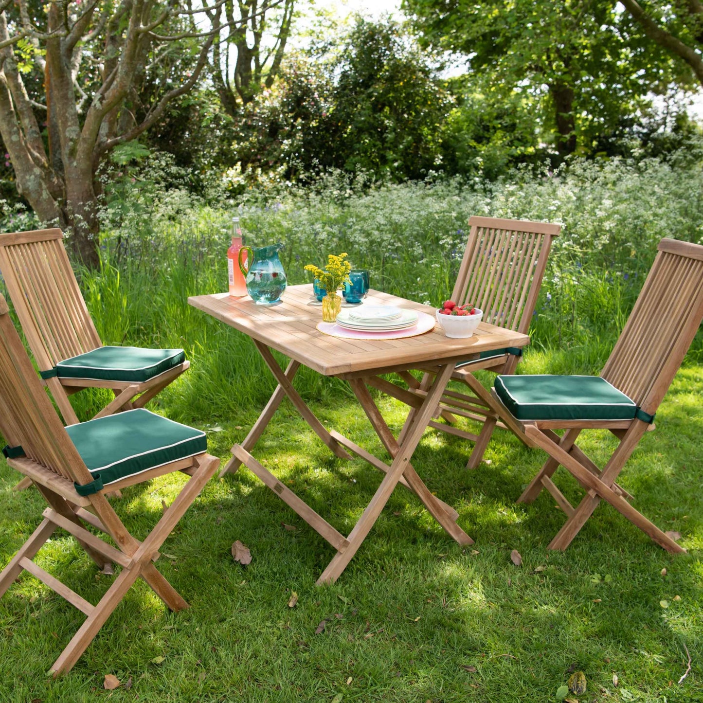 Teak folding dining chairs with green cushions, set at angles around teak folding rectangular dining table with green jug and glasses, in sunny garden.