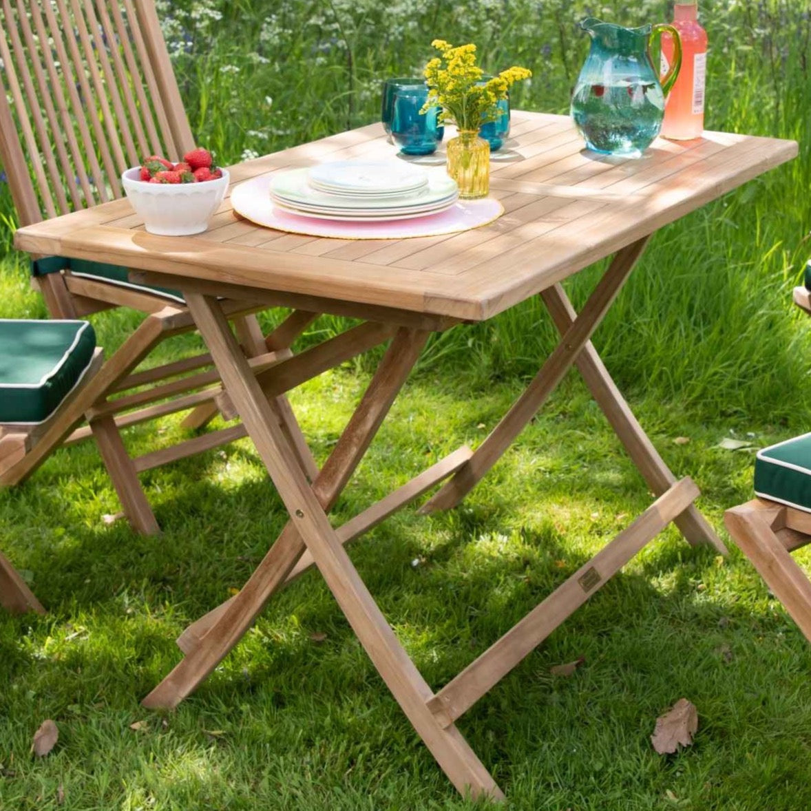 Teak folding rectangular table with teak chairs around with green seat cushions in a garden. On the table is a jug, glasses, fruit and plates. 