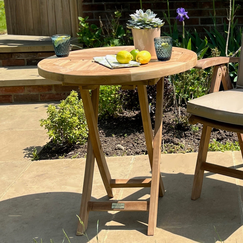 
                  
                    A small round folding table made of teak, set on a patio with folding legs, surrounded by garden elements. The table is accompanied by a teak chair with a cushion.
                  
                