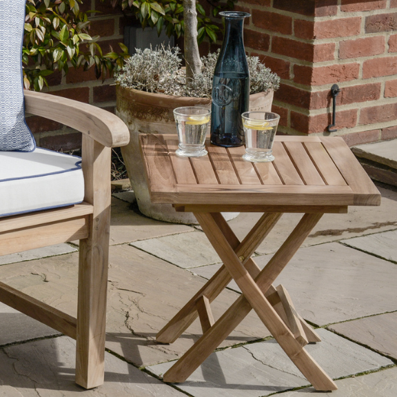 Small, folding teak coffee table on a patio, next to a  teak chair with cushions. On the table are two glasses of water and a bottle.  