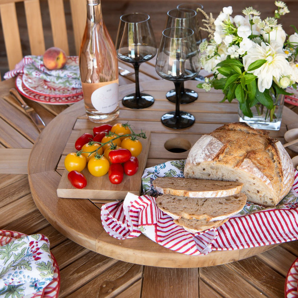 
                  
                    Round teak lazy susan turntable set with food, drinks and flowers, on a teak dining table with place settings.
                  
                