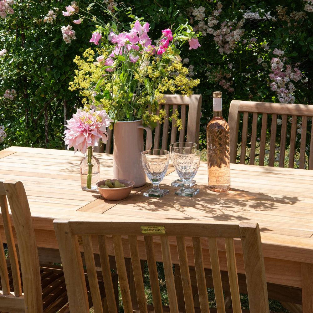 Outdoor setting with a teak rectangular table, teak chairs, and floral arrangements against a backdrop of greenery and flowers.