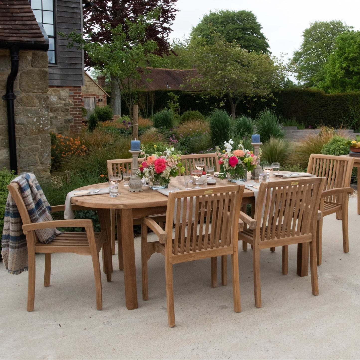 Teak outdoor dining set with oval table and six teak chairs on patio with flowers and place settings. 