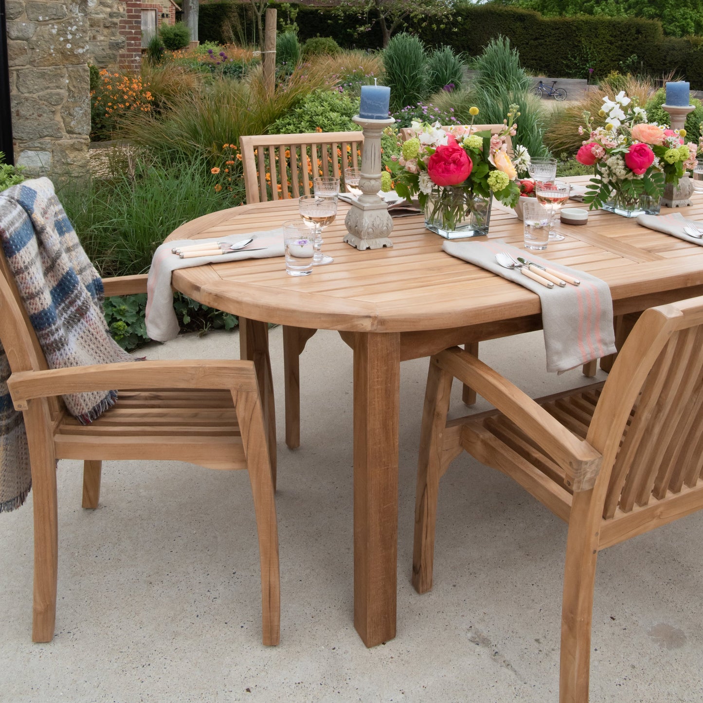 Teak outdoor dining set with oval teak table and chairs, with flowers and place settings, on a patio. 