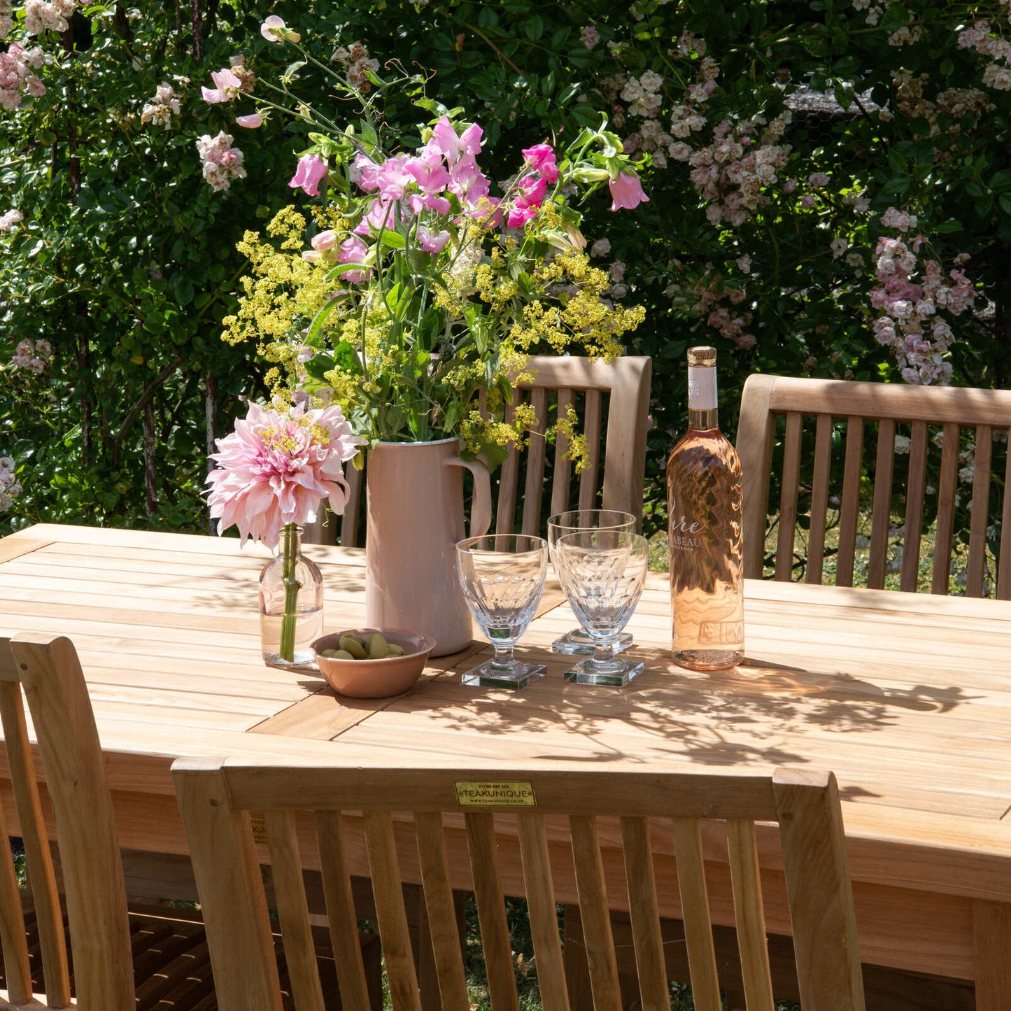 Outdoor setting with a teak wooden table, teak wooden chairs, and floral arrangements against a backdrop of greenery and flowers.