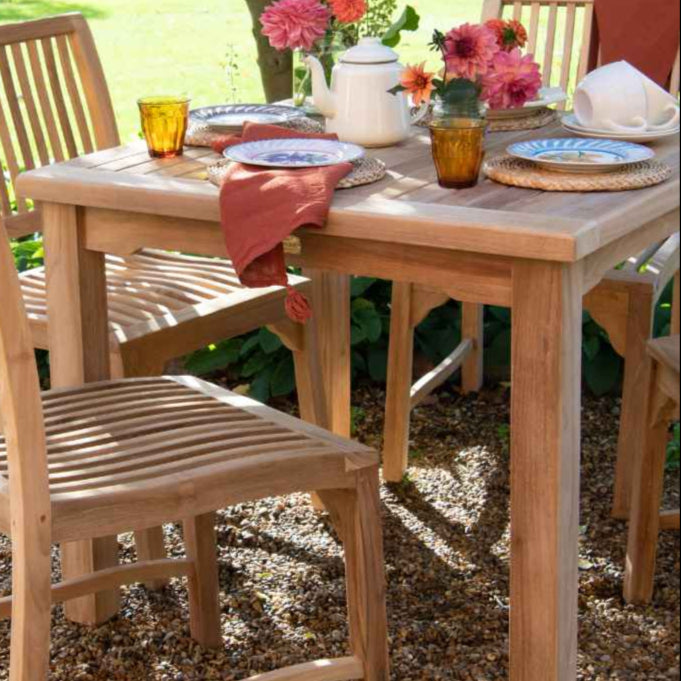 A square teak dining table with a slatted tabletop, central bar, and parasol hole, set on a gravel surface with chairs around it, table is arranged with plates, cutlery, and a vase of flowers.