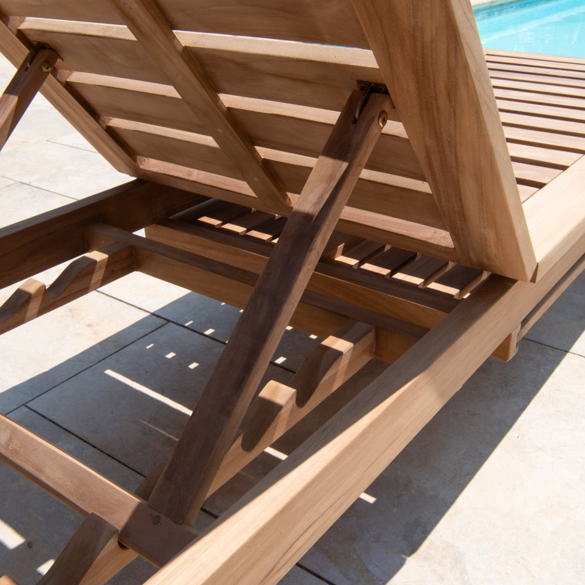 Close up of back of teak reclining chair to display teak wood struts and supports, on stone patio with pool in background 