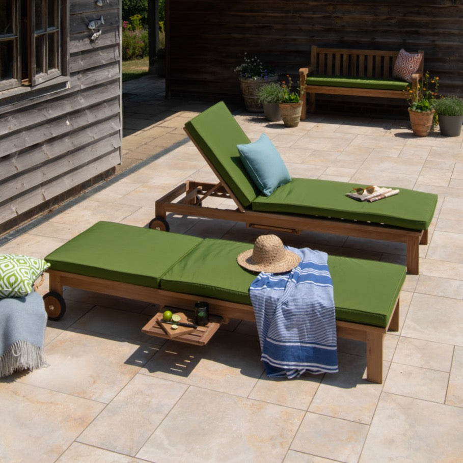 Two green cushioned teak wood loungers with chrome hardware, featuring a hidden drinks table, positioned on a patio with a wooden house backdrop and surrounded by garden decor.