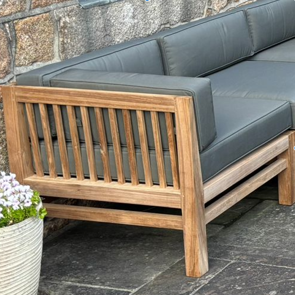 
                  
                    A modular teak sofa corner unit with gray cushions, placed on a flagstone surface and against a stone wall, including a ceramic planter to the side.
                  
                