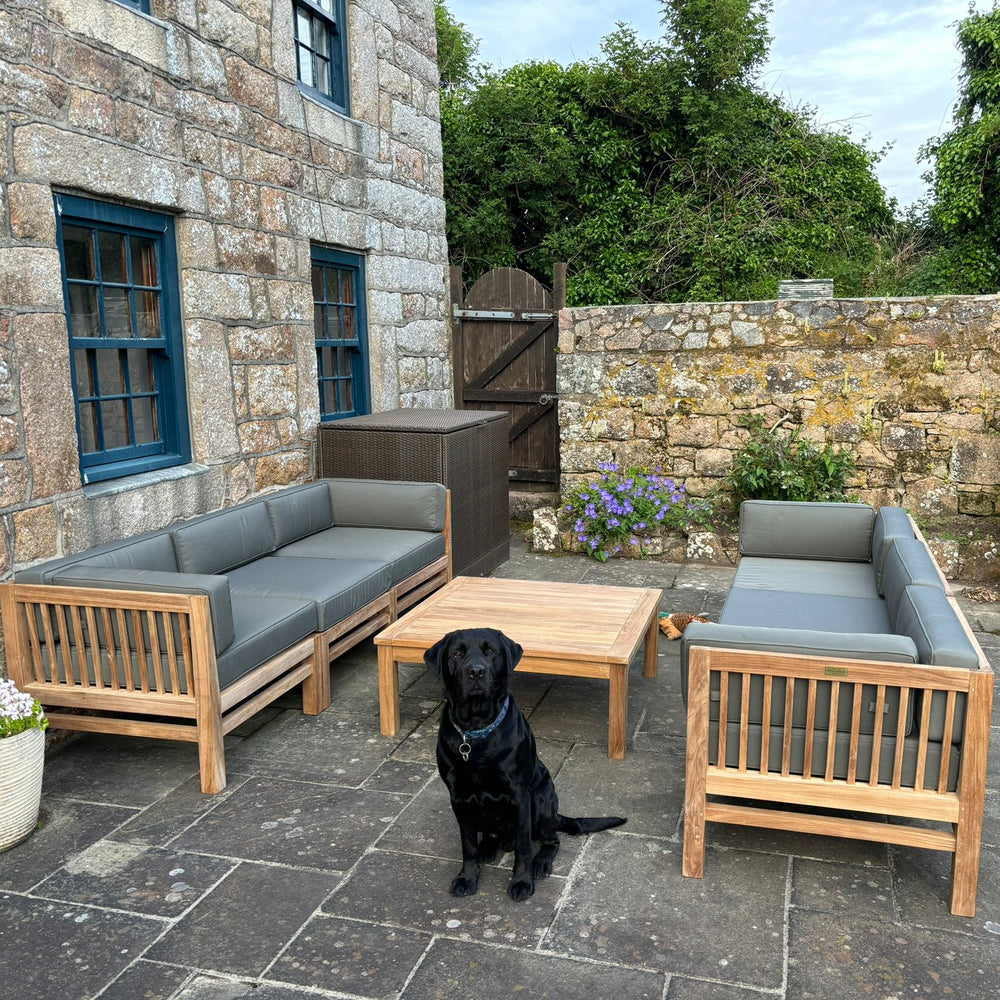 
                  
                    Patio with two teak outdoor sofas facing each other, with gray cushions and a low teak coffee table in between. Black dog sits looking at camera, in a Stone building setting
                  
                