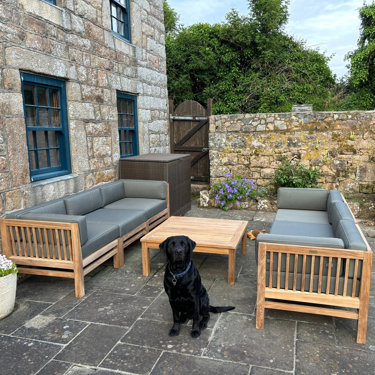 Patio with two teak outdoor sofas facing each other, with gray cushions and a low teak coffee table in between. Black dog sits looking at camera, in a Stone building setting