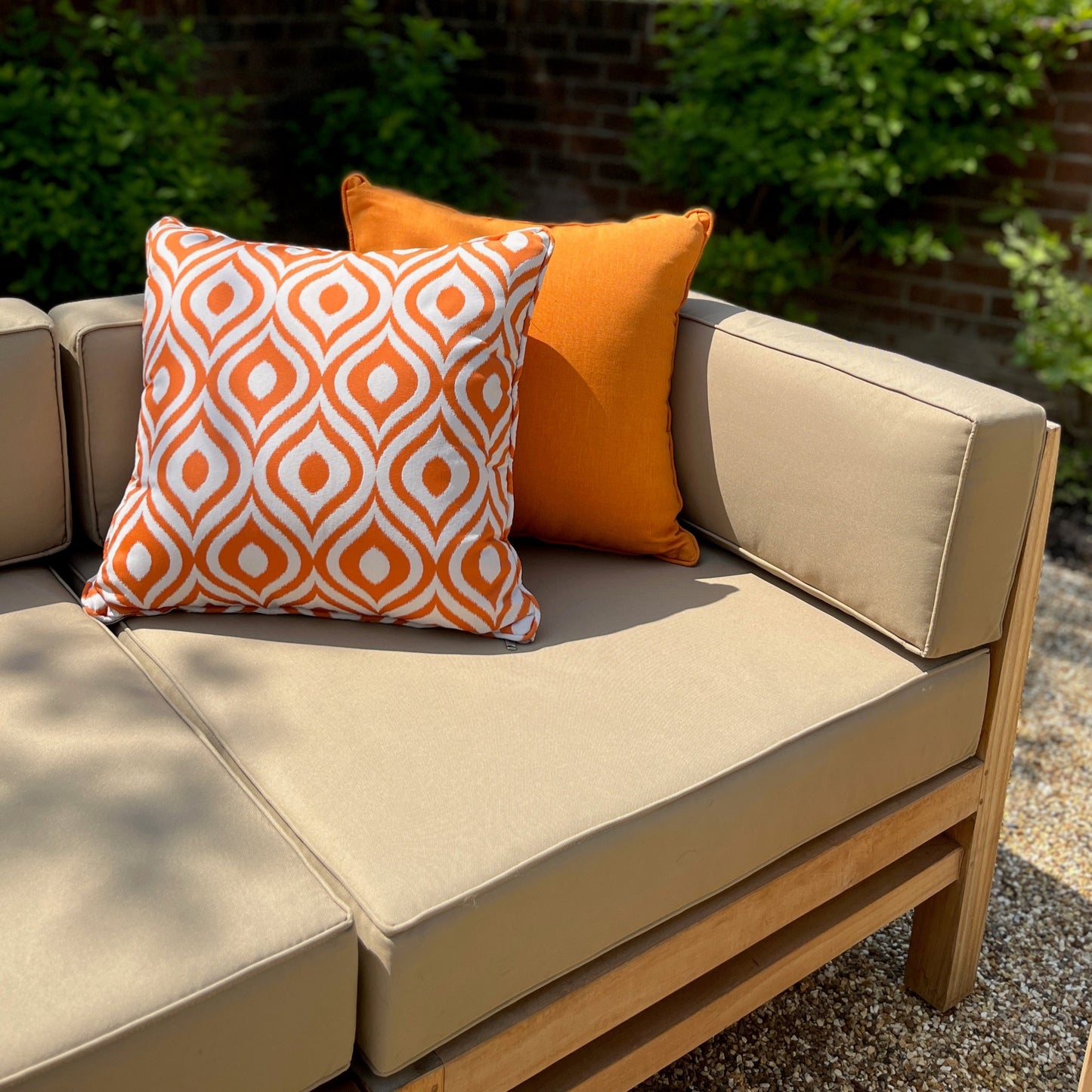 Corner section of teak outdoor sofa with oatmeal coloured seat, back and arm cushions and orange patterned scatter cushions, on gravel patio with brick wall behind 