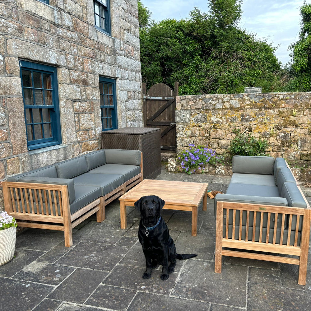 
                  
                    Two teak outdoor sofas with grey cushions, with low teak coffee table in between, on patio in front of stone building with black dog sitting.
                  
                