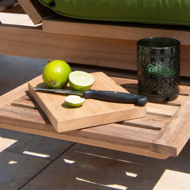 A small rectangular teak mini serving board placed on a teak table with a sliced lemon and a knife on top of it, next to a black decorative cup.