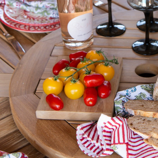 Mini teak serving board holding tomatoes, on top of round teak table with glasses, wine bottle and bread.  